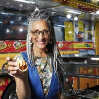 Host Carla Hall posing out front of Taco Tamix food truck on Chasing Flavor, season 1.